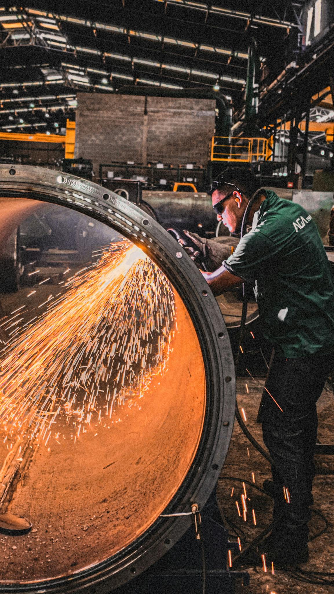 Worker grinding metal pipe in factory with sparks flying.